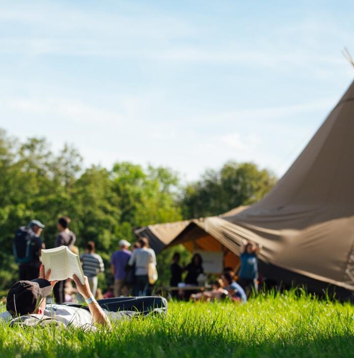 image of man lying down on the gras reading a book, with other festival goes in the background and a tent.