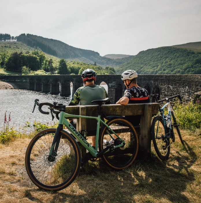 Two cyclists sat on a bench by a multi-arched bridge over a lake.