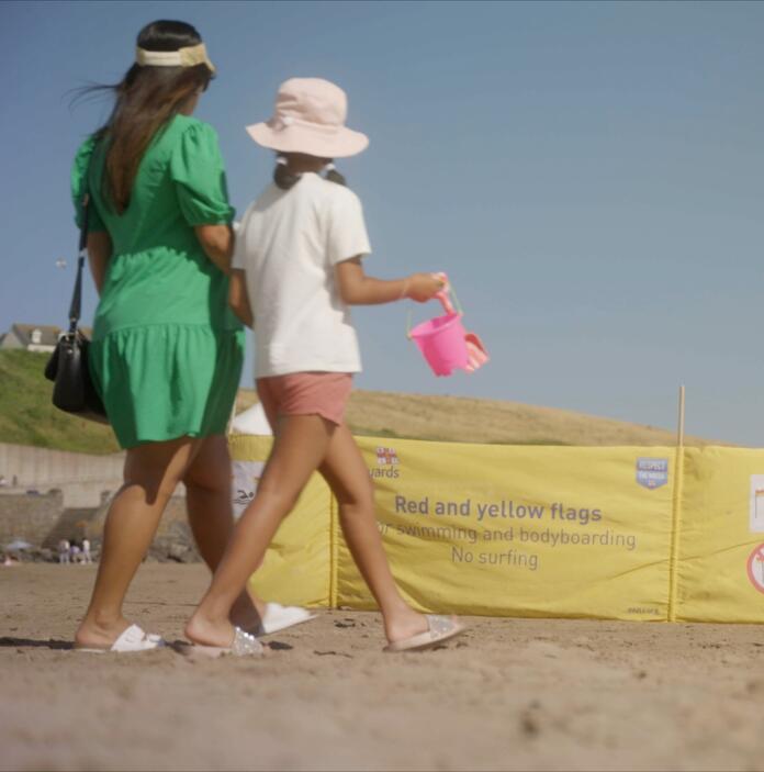 Two people walking on a beach with a yellow beach safety sign in the background.
