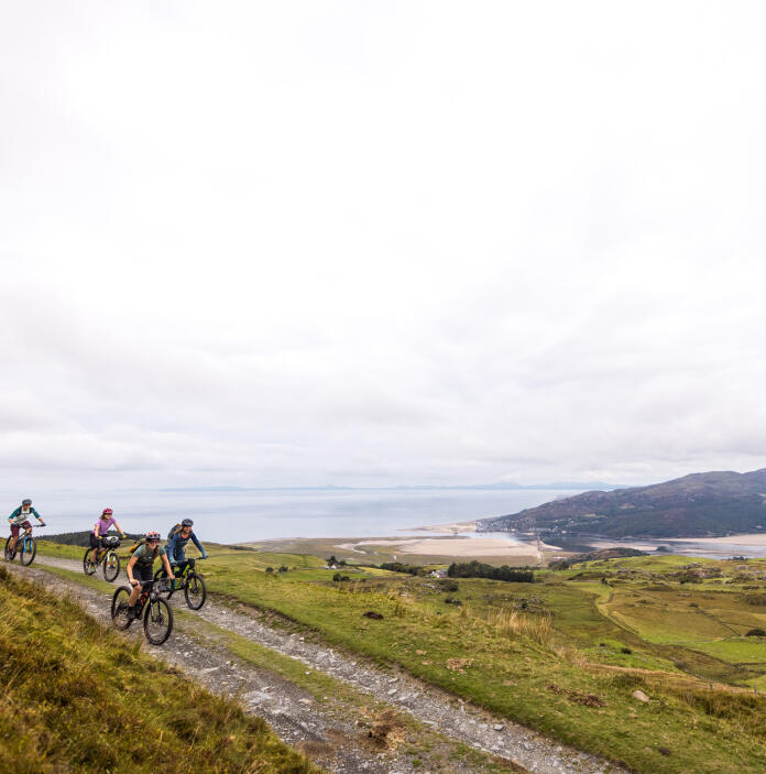 Four cyclists riding mountain bikes riding a rough mountain top track, with views over an estuary. 