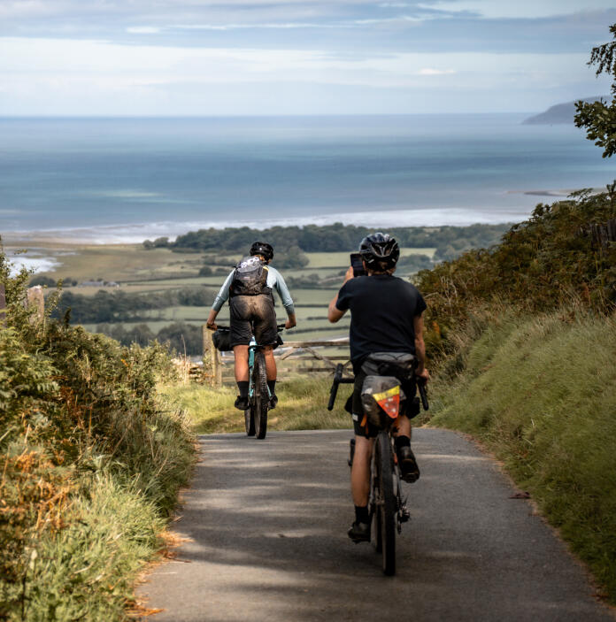 Two mountain bikers riding down a narrow road with views towards the coast.
