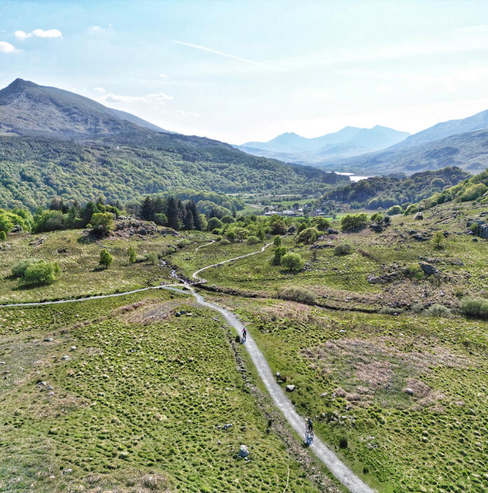 Aerial view of mountain bikers on a narrow road in a wide valley, cycling towards big mountains.