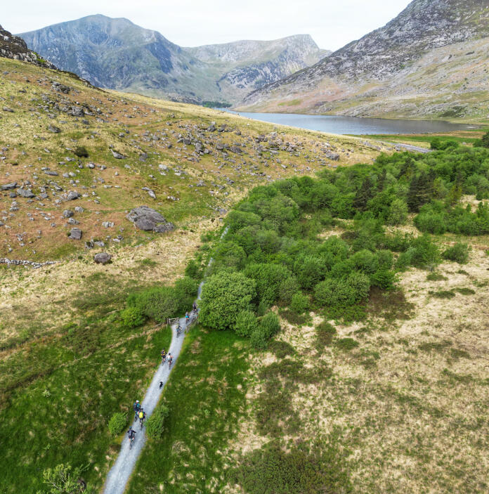 Aerial view of cyclists riding along a narrow road towards a lake.