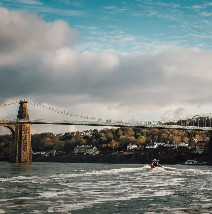 Suspension bridge with houses and trees in the distance, and a rib boat on the Menai Strait