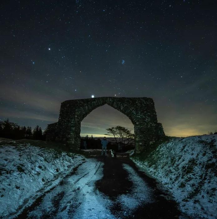 A stone built archway over a snowy narrow road at night. The stars are bright in thedark skies.