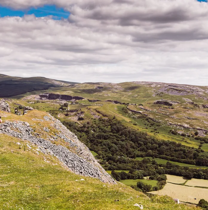 Two walkers on a hillside path overlooking a green valley strewn with rocks and woodland.