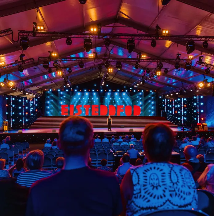 People watching performer on stage with large neon sign 'Eisteddfod'.