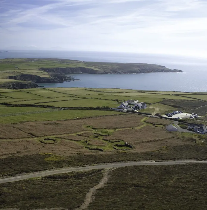 Aerial view of a rugged coastline with green fields and cliffs leading to a calm bay under a partly cloudy sky.