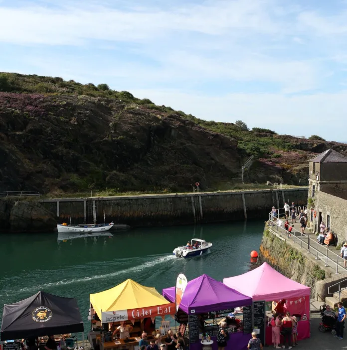 Outdoor market scene at a small harbor with colourful tents (yellow, pink, purple, black) set up along the waterfront. People are seated at wooden picnic tables near the stalls, and a small white boat is moored in the calm water. Stone buildings and a sloping green hillside form the backdrop under a partly cloudy sky.