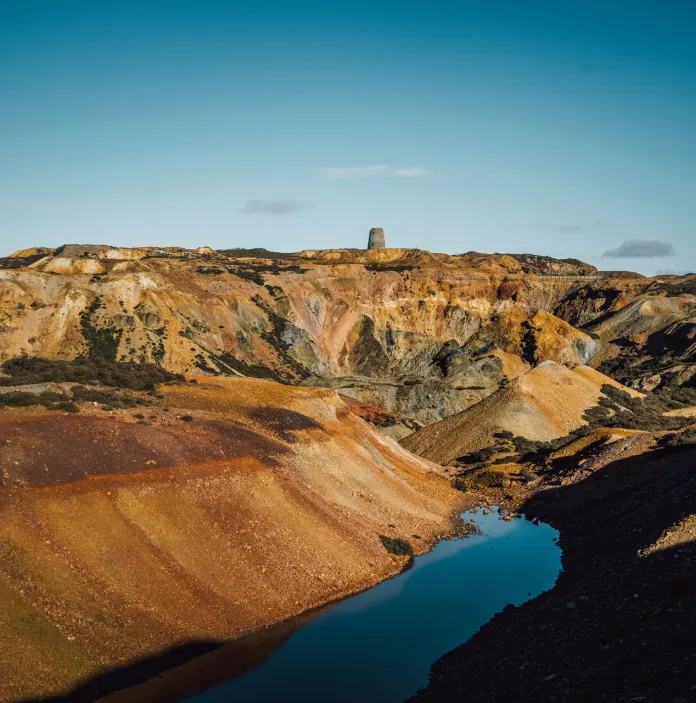 A vivid view of Parys Mountain’s colourful open‑cast landscape, showing rugged yellow, orange and brown slopes surrounding a deep blue water pool, with the historic windmill tower visible on the ridge beneath a bright blue sky.