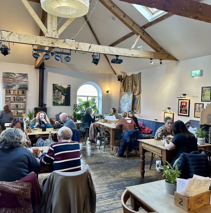 Spacious café or hall with wooden beams, filled with people sitting at tables and chatting.