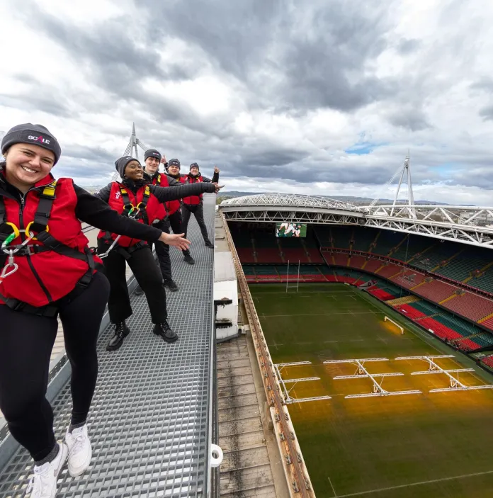 Person in a red jacket and helmet clipped to a safety line while walking along a high metal walkway beside a large dam, with water visible below and cloudy sky above.
