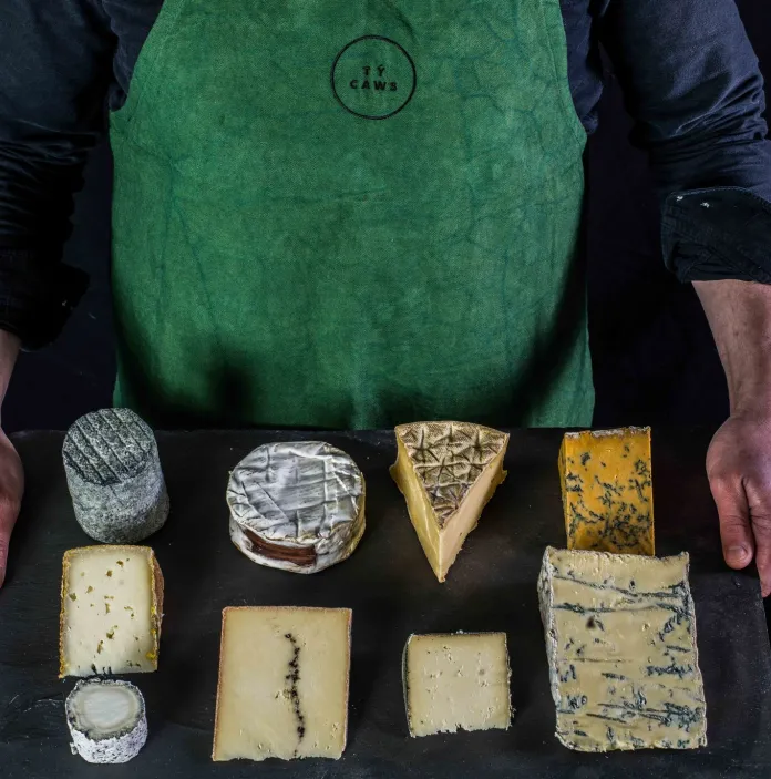 Selection of Welsh artisan cheeses arranged on a table, including wedges, rounds and blue cheeses, with a person standing behind the display wearing a green apron.