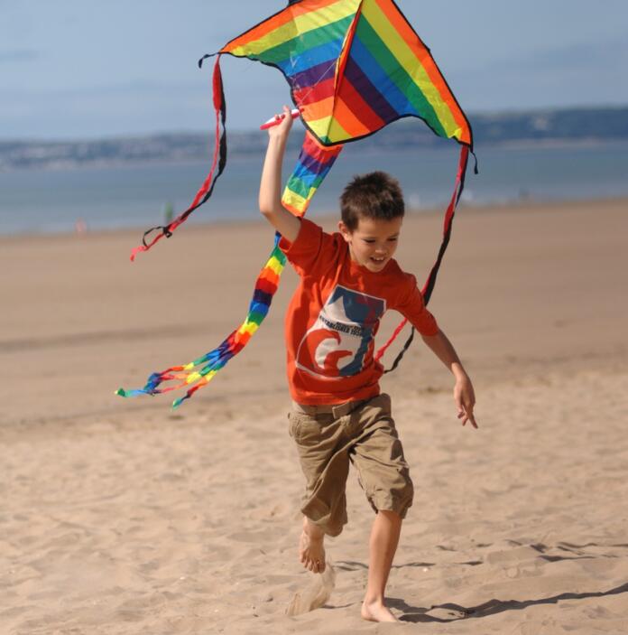 Boy running on a sandy beach with a big rainbow coloured kite