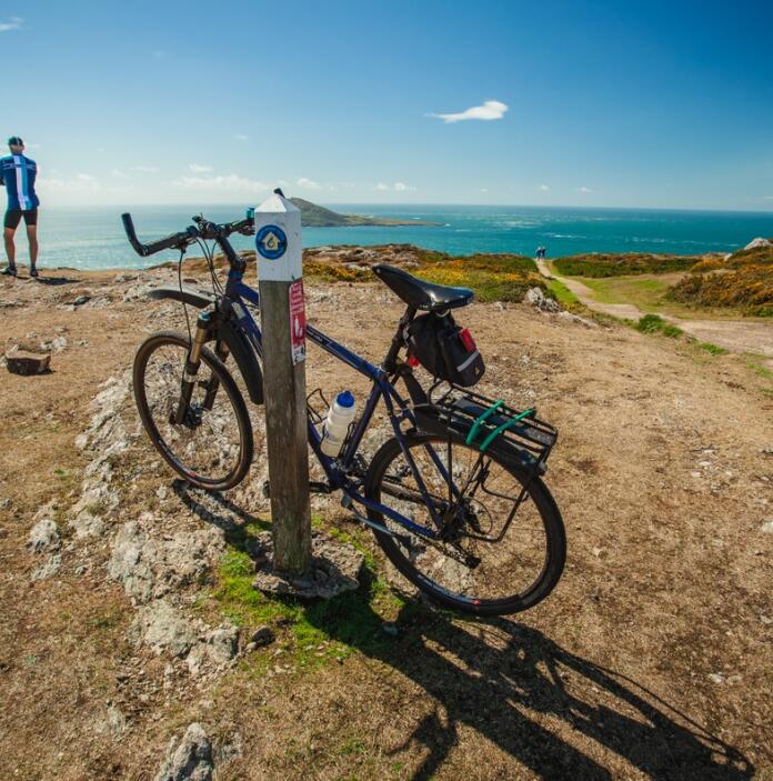 Ein Fahrrad, das an ein Schild des Wales Coast Path gelehnt ist, und ein Radfahrer im Hintergrund, der auf das Meer hinausschaut.