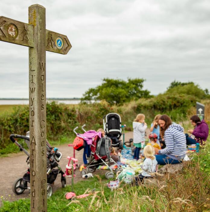 Eine Gruppe Eltern mit Kleinkindern und Kinderwagen macht eine Pause an einem Schild des Wales Coast Path.