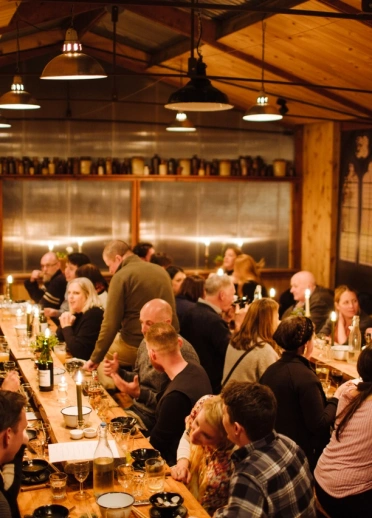 Lots of people on long dining tables in a wooden barn with glazed windows.