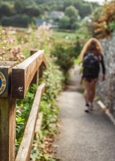 Das Schild des Wales Coast Path an einem Geländer mit einer Frau, die einen asphaltierten Weg entlang geht.