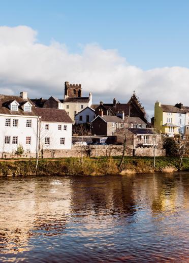 Fluss mit Häusern am Flussufer und die Spitze der Brecon Cathedral in der Ferne.
