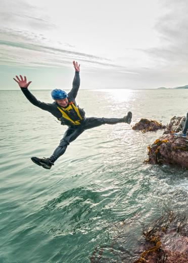Group coasteering on rocks with one member star jumping into the sea.