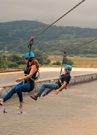 Two people zip wiring over a surfing lagoon with the countryside in the background.