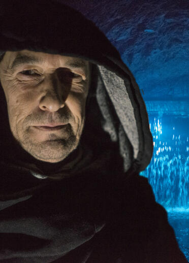 man with hood in foreground with cave and water light up in blue in the background 