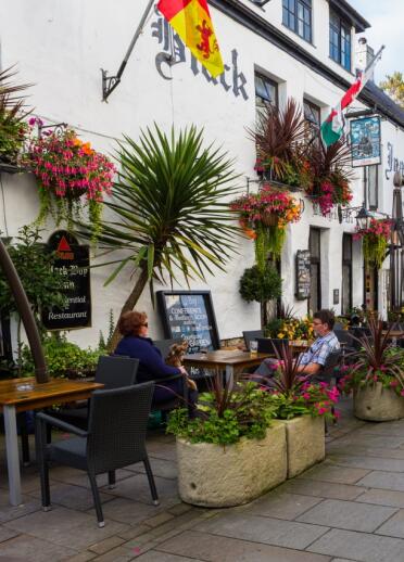 people sat at tables outside a pub with raised flower beds and a menu board.