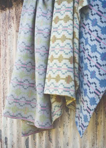 Image of four Welsh wool blankets with the same pattern but different colours hanging on the side of a hut