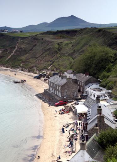 Nefyn golf course in teh distance with green mountain leading onto beach with pub and houses, people on teh beach and red van parked next to the inn.