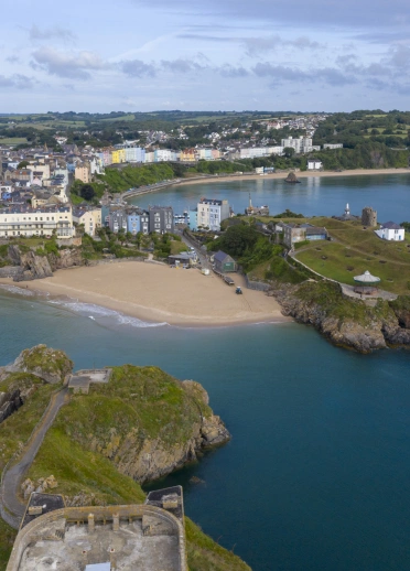 A small sandy cove, with a green hill on one side and colourful houses behind.