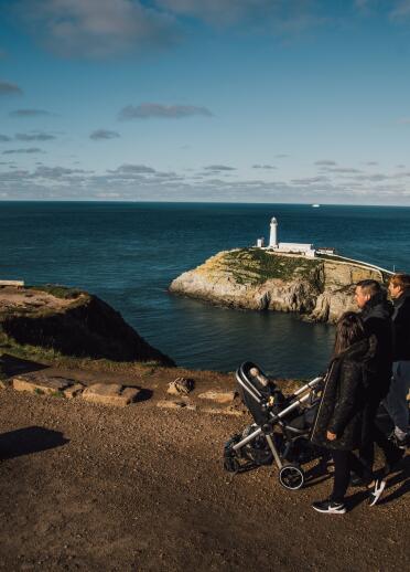two men and a woman walking with a pushchair, with lighthouse and coast in background.