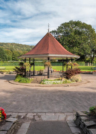 A bandstand in the middle of a green leafy park with flowers and wooden benches surrounding it. The sky is blue. 