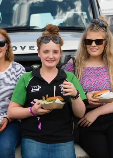 Three women sitting down enjoying burgers. A silver Land Rover and a caravan are in the background.
