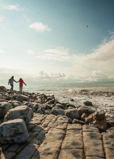 Zwei Personen wandern an einem Steinstrand mit Klippen im Hintergrund.