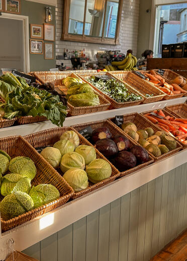 Verkaufsraum eines Lebensmittelgeschäfts mit Kisten mit Gemüse wie Lauch, Kohl, Karotten und Kartoffeln im Vordergrund.
