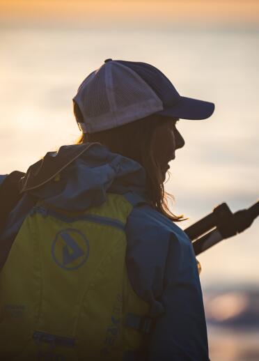 Back shot of paddleboarder wearing a peaked cap and yellow buoyancy aid, holding a paddle, silhouetted against a reflection of the sun on the sea.