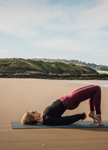 Person in a yoga pose on a beach with grassy dunes and fields in the distance