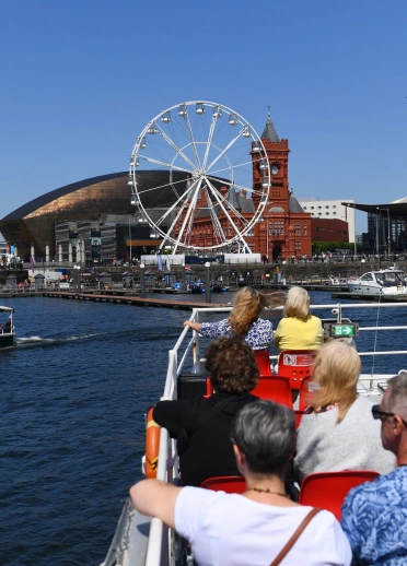 People on a boat in a bay, looking at a marina, a Ferris wheel and architecturally striking buildings.