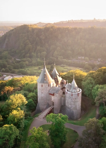 Aerial shot of a fairy tale style castle with pointy turrets and a drawbridge, set amongst the trees of a woodland. 