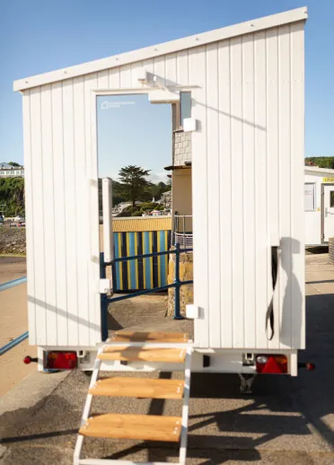 The exterior of a mobile sauna with white walls and wooden steps up to a mirrored door.