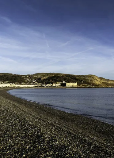 A wide, pebbly beach looking towards a pier and a hill.