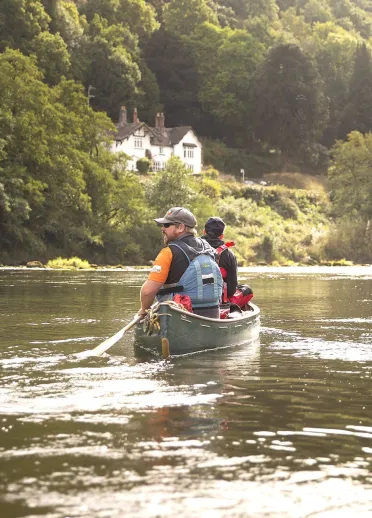 Two people canoeing on a wide, reflective river, surrounded by dense woodland, with a white house visible on the riverbank ahead.