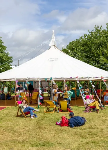 Large white marquee tent on grass at outdoor festival, with people sitting on deckchairs and blankets, surrounded by trees under a partly cloudy sky.