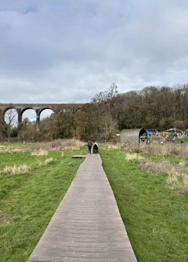 A wooden broadwalk in a country park, with trees and a railway viaduct in the background.