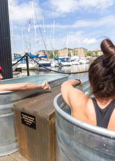 Two people sitting in separate outdoor ice baths beside a marina, with boats and apartment buildings in the background and a sign reading “Please shower before using ice baths”.