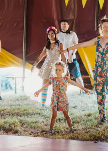 Festival-goers of all ages take part in a family dance session inside a decorated festival tent, with a child standing at the front of the group.