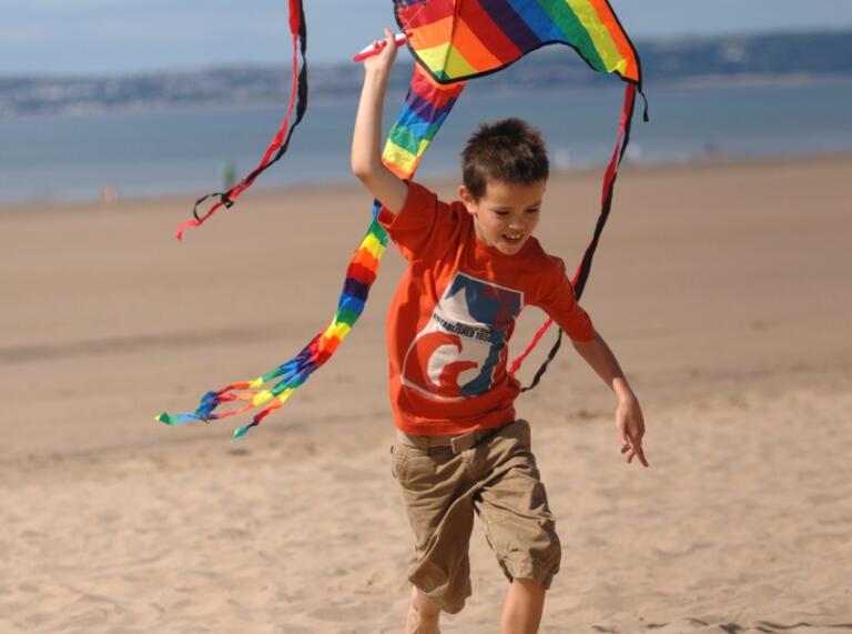 Junge, der mit einem großen regenbogenfarbenen Drachen über einen Sandstrand läuft.