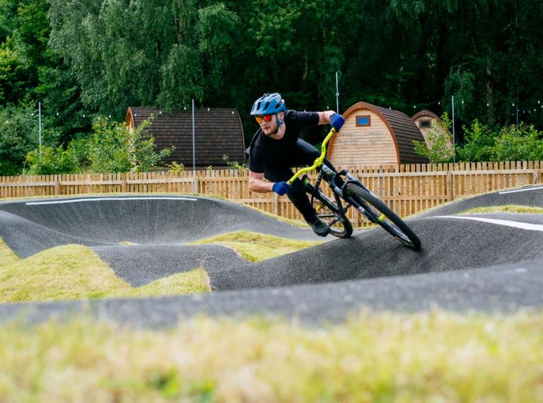 A mountain biker cycling around a pump track with glamping pods in the background.