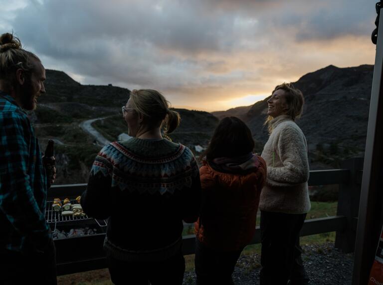 Four friends laughing and socialising on the balcony of a glamping pod overlooking the slate landscape of Blaenau Ffestiniog.