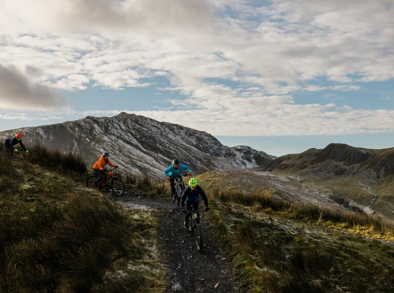 Four people on mountain bikes riding down a single track mountain trail with mountain scenery in the background.